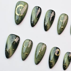 May include: Olive green press-on nails with gold celestial designs. Each nail features a gold crescent moon, stars, and small dots. The nails have a glossy finish and are arranged on a white surface. A set of stylish, ready-to-wear artificial nails.