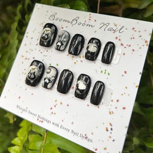 This nail set features a playful black base adorned with whimsical silver characters and patterns, adding a fun and mysterious vibe. Perfect for those who love unique, eye-catching designs with a touch of charm.