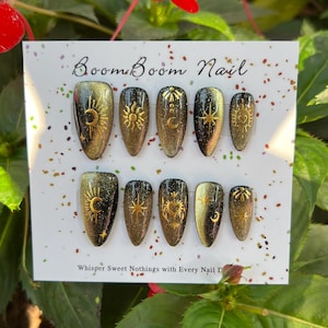 K&ouml;nnte beinhalten: Ein Set aus zehn gold-schwarzen Kunstn&auml;geln mit Himmelsmotiven, darunter Sonnen, Monde und Sterne. Die N&auml;gel sind auf einer wei&szlig;en Karte mit dem Text "Boom Boom Nail" und "Whisper Sweet Nothings with Every Nail D" angeordnet.