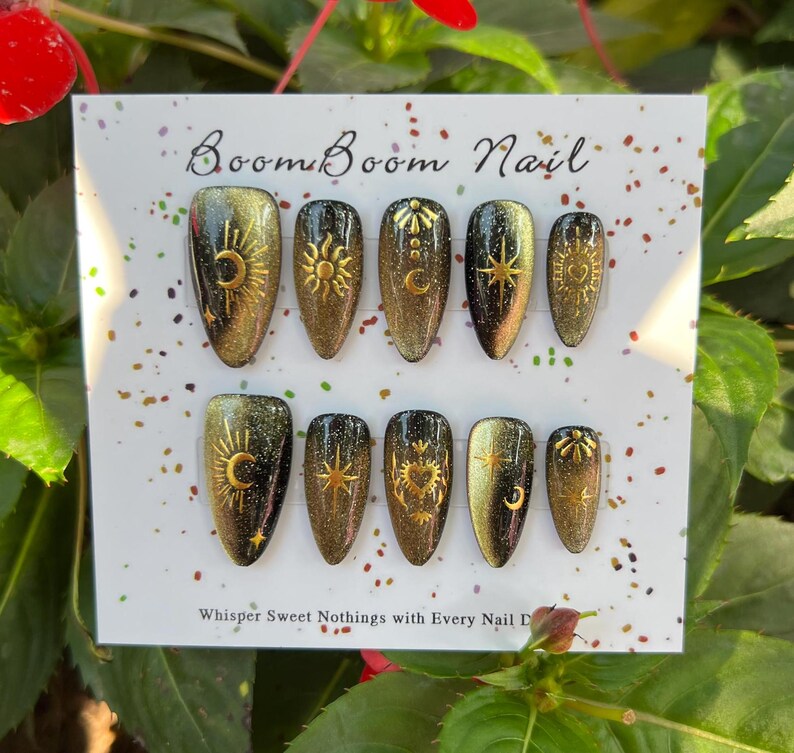 K&ouml;nnte beinhalten: Ein Set aus zehn gold-schwarzen Kunstn&auml;geln mit Himmelsmotiven, darunter Sonnen, Monde und Sterne. Die N&auml;gel sind auf einer wei&szlig;en Karte mit dem Text "Boom Boom Nail" und "Whisper Sweet Nothings with Every Nail D" angeordnet.