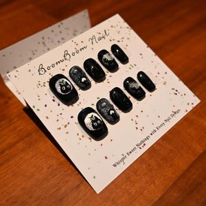 This nail set features a playful black base adorned with whimsical silver characters and patterns, adding a fun and mysterious vibe. Perfect for those who love unique, eye-catching designs with a touch of charm.