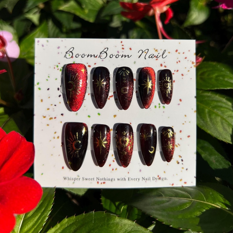 Puede incluir: Un conjunto de diez u&ntilde;as postizas rojas y negras con dise&ntilde;os celestiales dorados. Las u&ntilde;as est&aacute;n dispuestas en una tarjeta blanca con el texto "Boom Boom Nail" y "Whisper Sweet Nothings with Every Nail Design."
