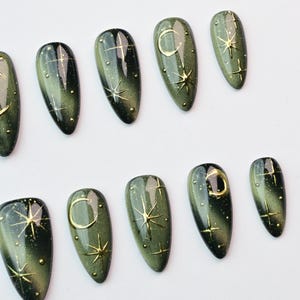 May include: Set of olive green press-on nails with gold celestial designs. Each nail features a gradient effect, with gold stars, moons, and dots. The nails are oval-shaped and have a glossy finish.