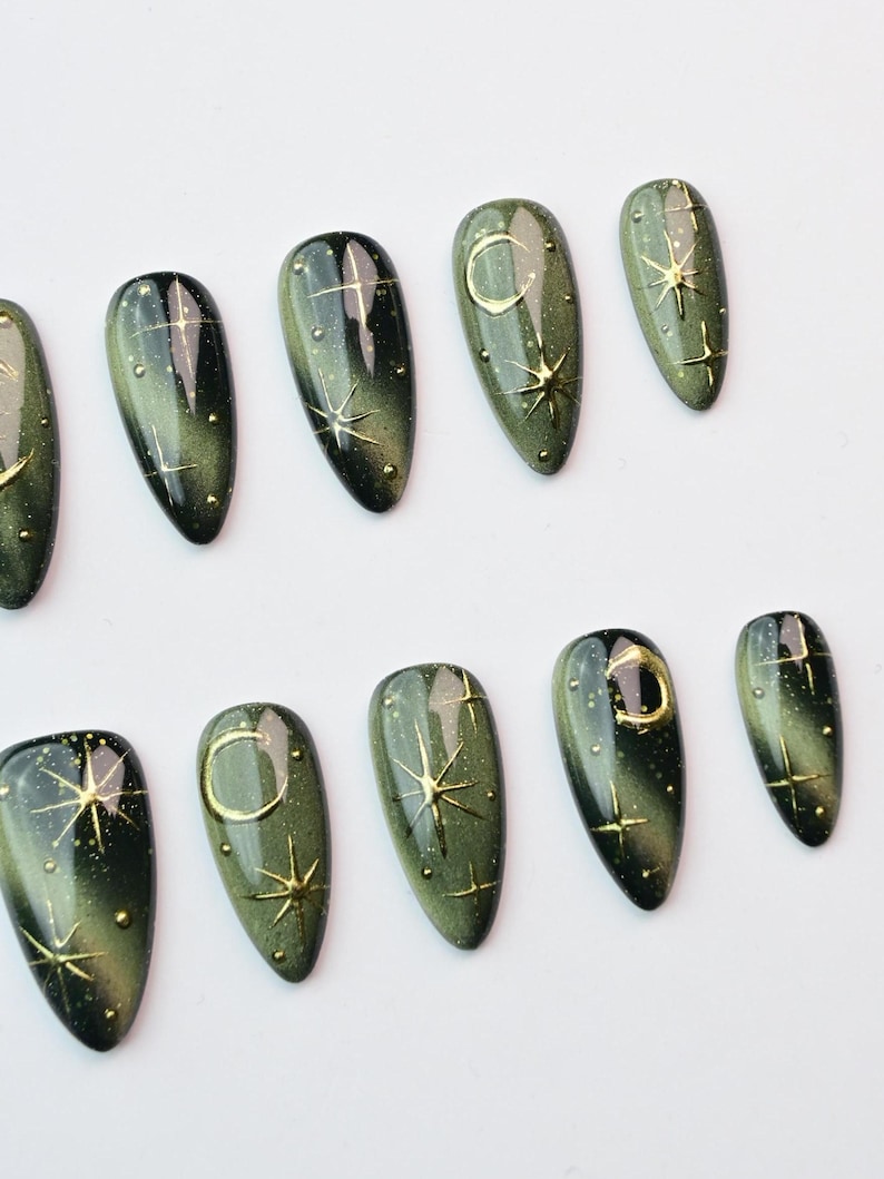 May include: Set of olive green press-on nails with gold celestial designs. Each nail features a gradient effect, with gold stars, moons, and dots. The nails are oval-shaped and have a glossy finish.