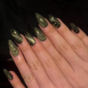 May include: A set of long, almond-shaped artificial nails. The nails are painted a dark olive green with a cat-eye effect. Gold celestial designs, including stars and crescent moons, adorn the nails. The nails are on a person's hand.