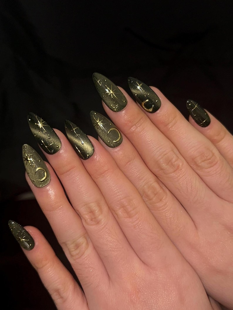 May include: A set of long, almond-shaped artificial nails. The nails are painted a dark olive green with a cat-eye effect. Gold celestial designs, including stars and crescent moons, adorn the nails. The nails are on a person's hand.