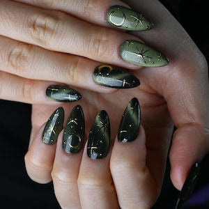 May include: Close-up of a hand with long, almond-shaped nails. The nails are painted a dark olive green with a cat-eye effect. Gold accents include crescent moons and starburst designs. The nails are a stylish accessory.