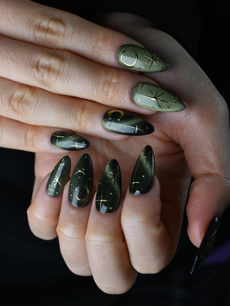 May include: Close-up of a hand with long, almond-shaped nails. The nails are painted a dark olive green with a cat-eye effect. Gold accents include crescent moons and starburst designs. The nails are a stylish accessory.