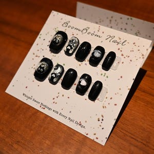 This nail set features a playful black base adorned with whimsical silver characters and patterns, adding a fun and mysterious vibe. Perfect for those who love unique, eye-catching designs with a touch of charm.