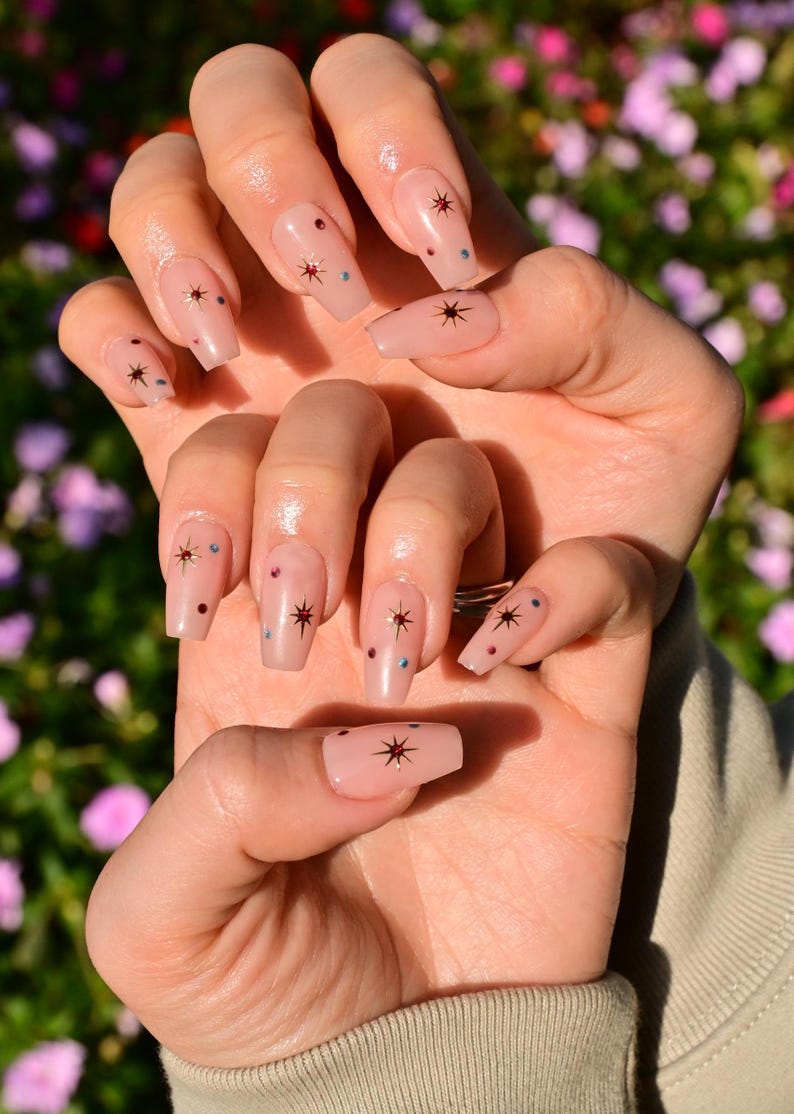 This nail set features a soft nude pink base adorned with delicate golden starbursts and colorful confetti details. Perfect for a playful yet elegant look that adds a touch of sparkle to any occasion.