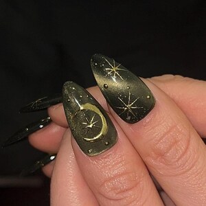 May include: Close-up of long, almond-shaped nails painted dark green with gold celestial designs. One nail features a gold crescent moon and star, while others display starburst patterns. The nails have a glossy finish.