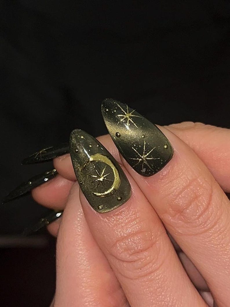 May include: Close-up of long, almond-shaped nails painted dark green with gold celestial designs. One nail features a gold crescent moon and star, while others display starburst patterns. The nails have a glossy finish.