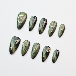 May include: Set of ten olive green press-on nails with a glossy finish. Each nail features a gold celestial design, including moons and stars. The nails are arranged on a white surface, showcasing their oval shape and intricate details.