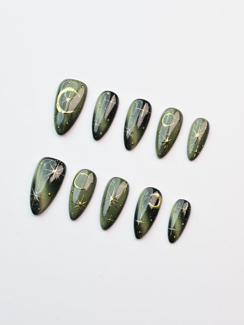 May include: Set of ten olive green press-on nails with a glossy finish. Each nail features a gold celestial design, including moons and stars. The nails are arranged on a white surface, showcasing their oval shape and intricate details.