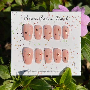 This nail set features a soft nude pink base adorned with delicate golden starbursts and colorful confetti details. Perfect for a playful yet elegant look that adds a touch of sparkle to any occasion.