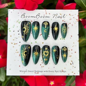 K&ouml;nnte beinhalten: Ein Set von Press-On-N&auml;geln mit einem schwarzen und smaragdgr&uuml;nen Glitzerdesign. Jeder Nagel zeigt goldene Himmelsmotive, darunter Monde, Sterne und Herzen. Auf der Karte steht "Boom Boom Nail" und "Whisper Sweet Nothings with Every Nail Design."