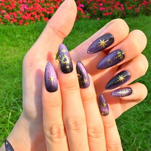 This nail design features a deep purple to black gradient base, adorned with gold celestial elements like the moon, sun, and stars, creating a luxurious and mystical vibe.