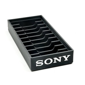 May include: A black rectangular tray with multiple slots and curved cutouts. The tray has the white "SONY" logo on the front. The tray is designed to hold small items.