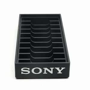 May include: A black plastic organizer with multiple slots and a curved central section. The word "SONY" is printed in white on the front. The organizer is designed to hold items and is set against a white background.