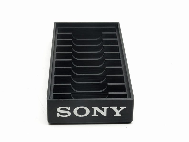 May include: A black plastic organizer with multiple slots and a curved central section. The word "SONY" is printed in white on the front. The organizer is designed to hold items and is set against a white background.