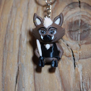 Könnte beinhalten: Ein brauner und weißer Cartoon-Wildschwein-Schlüsselanhänger mit einem schwarzen Shirt und einem weißen Stock. Das Wildschwein hat eine schwarze Nase und große braune Augen.