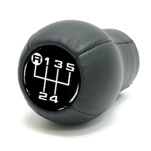 May include: A black gear shift knob with a white shift pattern diagram. The diagram shows the gear positions: R, 1, 2, 3, 4, and 5. The knob is made of a smooth, dark material and is set against a white background.