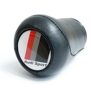 Puede incluir: Pomo de palanca de cambios de cuero negro con el logotipo de Audi Sport. El logotipo presenta rayas rojas, grises y negras sobre un fondo blanco. El pomo está diseñado para el cambio de marchas de un coche.