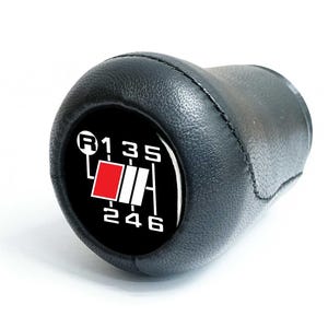 Op de afbeelding: Zwarte leren versnellingspookknop met een wit en rode versnellingspatroonindicator. De indicator toont de versnellingsposities: R, 1, 2, 3, 4, 5 en 6. De knop is voor een handgeschakelde versnellingsbak.