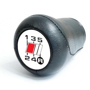 May include: Black leather gear stick knob with a white face showing the gear pattern. The pattern includes numbers 1, 3, 5, 2, 4, and R for reverse, with a red and white graphic. The knob is set against a white background.