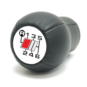 Puede incluir: Un pomo de palanca de cambios de cuero negro con una cara blanca que muestra el patrón de marchas. La cara tiene un logotipo rojo y plateado. El patrón de marchas incluye R, 1, 2, 3, 4, 5 y 6.