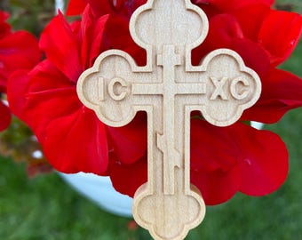 May include: A wooden Orthodox cross pendant with the Greek letters IC XC. The cross is light-colored and intricately carved, suspended by a gold-colored cord. The background features vibrant red flowers and green grass.