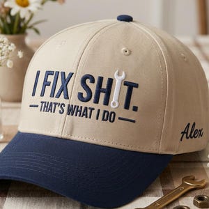 Puede incluir: Gorra de béisbol beige y azul marino con la frase bordada "I FIX SHIT. - THAT'S WHAT I DO -" y un gráfico de llave inglesa. El nombre "Alex" está bordado en el lateral.