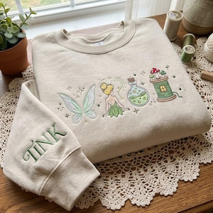 Könnte beinhalten: Beigefarbenes Sweatshirt mit gesticktem Feen-Design. Mit einer Fee, Flügeln, einer Zaubertrankflasche und Pilzen. Der Ärmel trägt die grüne Stickerei "TINK". Ein skurriles und magisches Kleidungsstück.