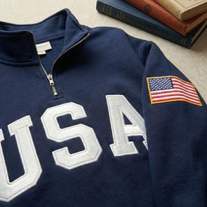 Peut inclure: Sweat-shirt bleu marine avec un col zippé et le mot "USA" brodé en blanc. Un écusson du drapeau américain est cousu sur la manche gauche. Le sweat-shirt est fait d'une matière douce et confortable.