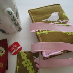 May include: A set of two fabric coasters with a bird and floral design in olive green, white, and pink, wrapped with a pink ribbon. A teacup and a tea bag are in the background.