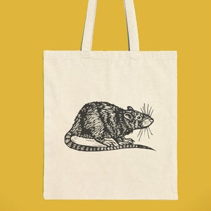 Könnte beinhalten: Eine weiße Canvas-Tragetasche mit schwarzen Henkeln und einer schwarz-weißen Illustration einer Ratte auf der Vorderseite.
