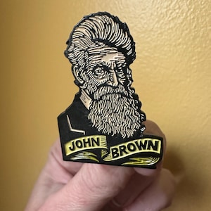 May include: A black and gold enamel pin featuring a portrait of John Brown with a long beard and the text "JOHN BROWN" on a banner. The pin is held against a yellow background.