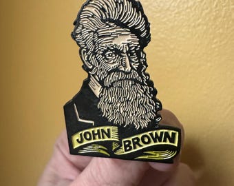 May include: A black and gold enamel pin featuring a portrait of John Brown with a long beard and the text "JOHN BROWN" on a banner. The pin is held against a yellow background.
