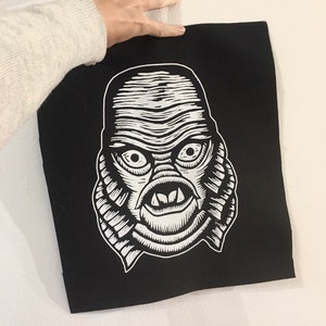 May include: Black fabric patch with a white graphic of the Creature from the Black Lagoon's face.