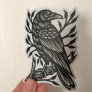 Peut inclure: Autocollant noir et blanc d'un corbeau perch&eacute; sur une branche avec des feuilles. Le corbeau est d&eacute;taill&eacute; avec des lignes et des ombres complexes.
