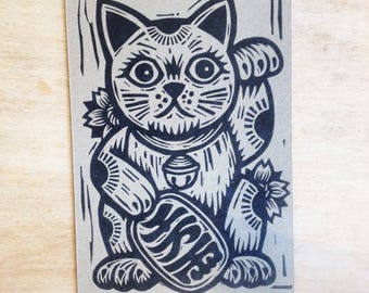 Op de afbeelding: Zwart-witte illustratie van een Japanse Maneki-Neko, of gelukkat, met een opgeheven poot. De kat houdt een munt in zijn andere poot en heeft een bel om zijn nek. De kat zit op een bruine achtergrond.