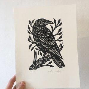 May include: Black and white linocut print of a crow perched on a branch. The print is signed and dated 2021.