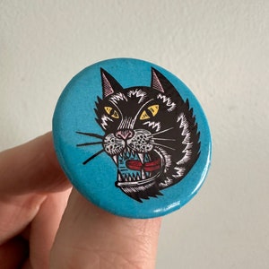 May include: A blue and black button with a cartoon image of a black cat with yellow eyes and a red tongue. The cat is snarling with its mouth open.