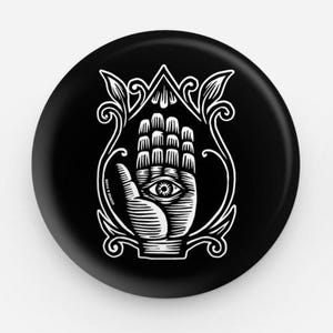 May include: Black and white button featuring a stylized hand with an eye in the palm, surrounded by ornate floral designs.