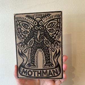 Può includere: Una stampa rettangolare su carta color marrone chiaro con un'illustrazione a stampa in inchiostro nero del Mothman. La creatura ha grandi ali con molteplici motivi a occhio, un corpo peloso e una lunga lingua. Un banner in basso riporta MOTHMAN.