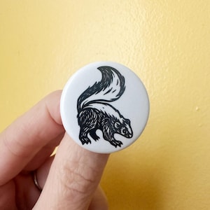 May include: A circular white pin-back button featuring a black ink-style illustration of a skunk with a raised tail. The design uses bold lines to depict the animal's fur texture and distinct markings, centered on the smooth, round surface.