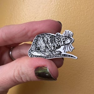 May include: A white enamel pin with a black and white line drawing of a rat. The rat is sitting on its hind legs with its tail curled around its body. The rat has a long, pointed snout and large, round ears.