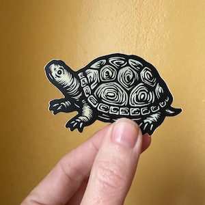 Pode incluir: Adesivo preto e branco de uma tartaruga com um design de casca detalhado. A tartaruga está andando para a direita.