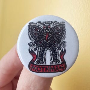 Może przedstawiać: Okrągła przypinka z czarno-białą ilustracją skrzydlatego stworzenia z wieloma oczami i czerwonym językiem. Pod stworzeniem znajduje się czerwony baner z napisem MOTHMAN wydrukowanym czarnymi wielkimi literami na białym tle.