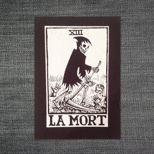 Puede incluir: Impresión de carta de tarot en blanco y negro con la Parca con una guadaña, el número XIII y las palabras "LA MORT". La ilustración representa a la Parca con una capa negra, caminando por un campo con manos y rostros.